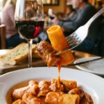 A close-up of a fork holding a piece of rigatoni and sausage dripping with creamy tomato sauce, with a glass of red wine in the background.