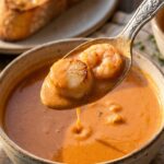 A close-up of a silver spoon lifting a bite of creamy orange bisque with a scallop and a piece of shrimp, with crusty bread in the background.