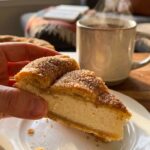 A close-up of a hand holding a square crescent cheesecake bar, showing the flaky cinnamon crust and thick cream cheese filling.