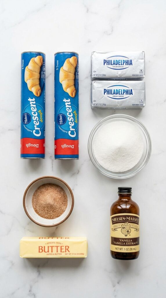 A flat lay showing tubes of crescent dough, cream cheese, sugar, butter, vanilla, and cinnamon on a marble board.