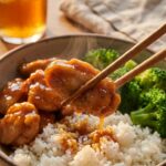 A close-up of chopsticks holding a piece of sticky bourbon chicken over a bowl of white rice and steamed broccoli.