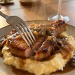A close-up of a fork cutting into a tender pork chop covered in gravy, served over a bed of mashed potatoes.