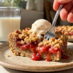 A close-up of a hand holding a warm strawberry oatmeal bar topped with melting vanilla ice cream, showing the gooey red center.