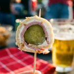 A close-up of a hand holding a pickle roll-up on a wooden toothpick with a tailgate party and beer in the background.