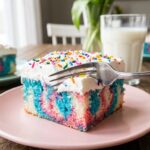 A close-up of a fork cutting into a slice of colorful tie-dye Easter poke cake on a pink plate.