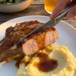 A close-up of a knife slicing into a juicy, glazed pork chop served over mashed potatoes, with the sauce dripping down the sides.