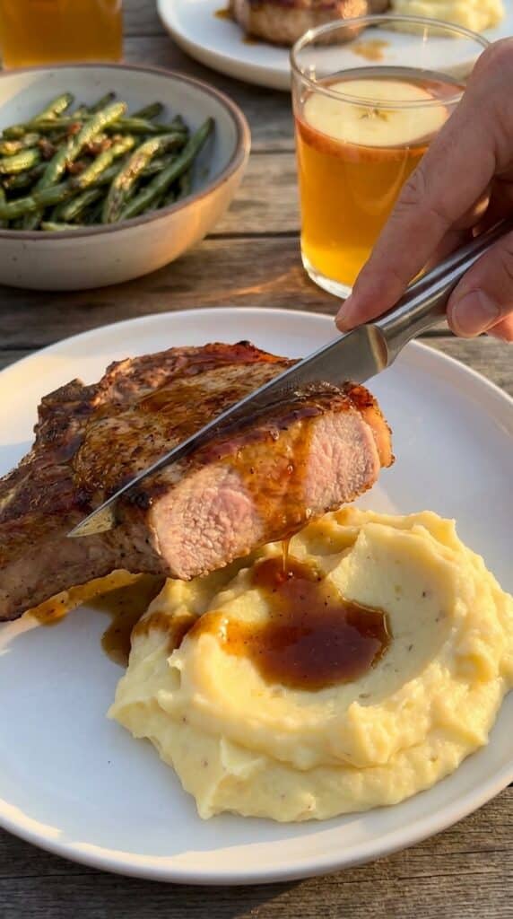 A close-up of a knife slicing into a juicy, glazed pork chop served over mashed potatoes, with the sauce dripping down the sides.