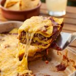 A close-up of a spatula lifting a layered slice of beef enchilada casserole with a dramatic cheese stretch, showing the tortilla and beef layers.
