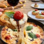 A close-up of a piece of toasted bread dipping into a hot Caprese dip, creating a gooey cheese pull with a tomato and basil on top.