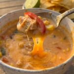 A close-up of a spoon lifting creamy chicken fajita soup with a dramatic melted cheese pull stretching back to the bowl.