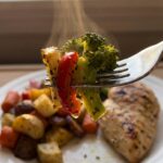 A close-up of a fork holding a bite of caramelized roasted broccoli and red bell pepper, with a plate of food in the blurred background.