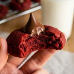 A close-up of a hand holding a red velvet blossom cookie with a bite taken out, showing the chewy center and chocolate kiss.
