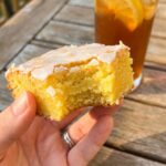 A close-up of a hand holding a lemon brownie with a bite taken out, showing a dense, fudgy interior, with iced tea in the background.