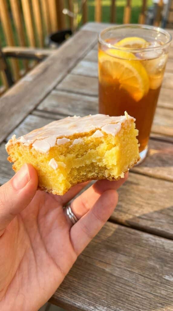 A close-up of a hand holding a lemon brownie with a bite taken out, showing a dense, fudgy interior, with iced tea in the background.