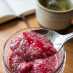 A close-up of a spoon scooping out a bite of thick chia pudding marbled with pink raspberry puree, with tea in the background.