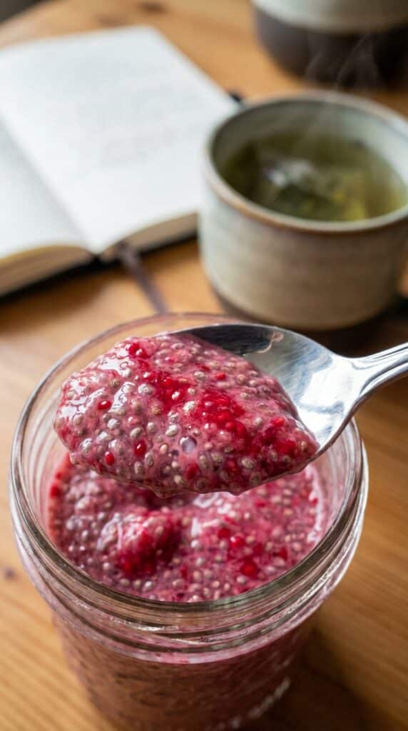 A close-up of a spoon scooping out a bite of thick chia pudding marbled with pink raspberry puree, with tea in the background.