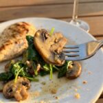 A close-up of a fork lifting a juicy, buttery mushroom and wilted spinach from a dinner plate, with grilled chicken blurred in the background.