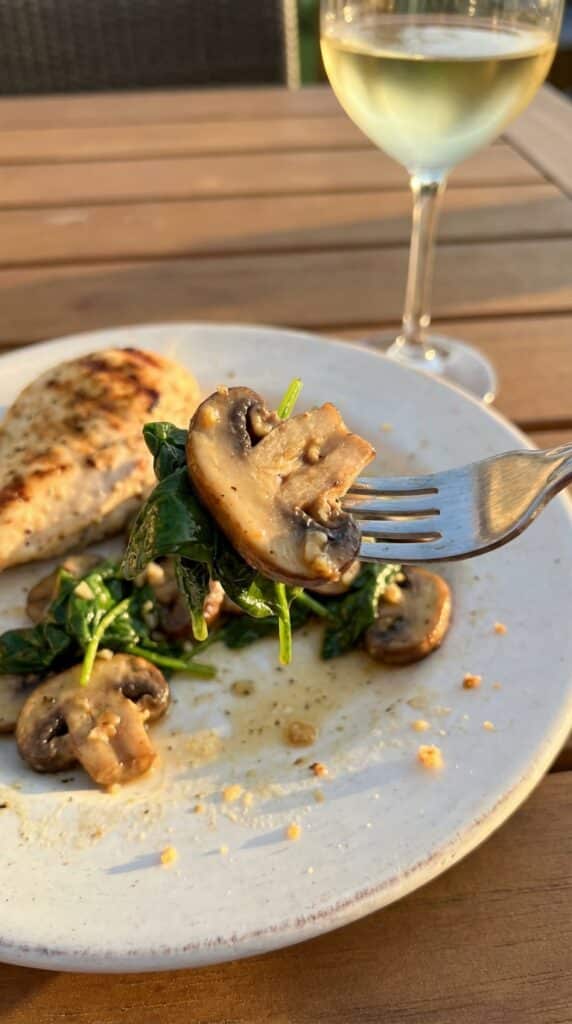 A close-up of a fork lifting a juicy, buttery mushroom and wilted spinach from a dinner plate, with grilled chicken blurred in the background.