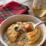 A close-up of a spoon lifting a bite of creamy chicken pot pie filling and a piece of flaky biscuit from a white bowl.