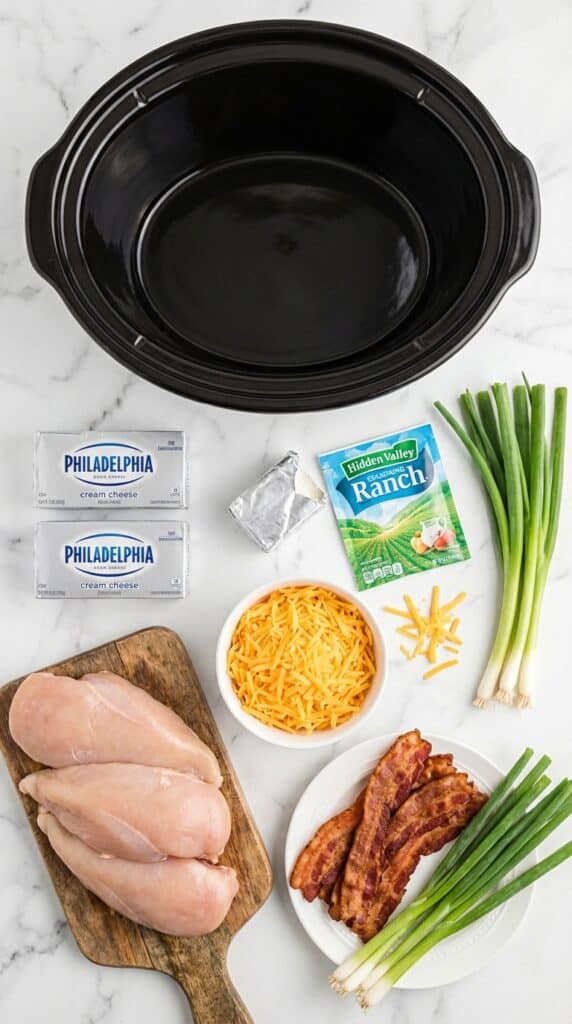 A flat lay showing raw chicken, cream cheese, ranch seasoning, cheddar cheese, cooked bacon, and green onions around a slow cooker insert.