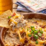 A close-up of a tortilla chip scooping up creamy, cheesy shredded chicken and bacon dip, creating a gooey cheese pull.