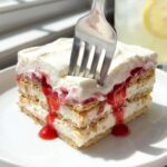 A close-up of a fork effortlessly cutting through a slice of layered strawberry icebox cake, showing the soft texture.