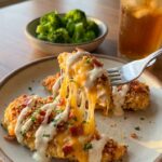 A close-up of a fork lifting a cheesy, bacon-topped chicken tender with a long cheese pull, served with a side of broccoli.