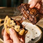 Two hands pulling apart a warm brookie, showing the gooey, melted chocolate stretching between the brownie and cookie halves.