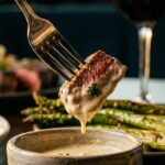 A close-up of a fork lifting a seared steak bite dripping with creamy parmesan sauce, with roasted asparagus in the background.