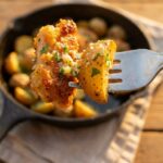 A close-up of a fork holding a piece of golden-brown garlic parmesan chicken and a roasted potato with the skillet in the background.