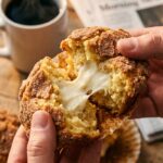 A close-up of two hands pulling apart a warm cinnamon muffin, showing the gooey cream cheese center stretching slightly.