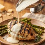 A close-up action shot of a silver pitcher pouring creamy white garlic parmesan sauce over a grilled stuffed chicken breast with grill marks.
