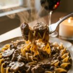 A close-up of a fork lifting a bite of creamy ground beef stroganoff and egg noodles from a plate, with steam rising.