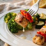 A close-up of a fork holding a piece of seared chicken and red bell pepper over a plate of healthy skillet vegetables.