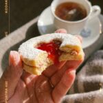 A close-up of a hand holding a heart jam cookie with a bite taken out, showing the jam filling, with a cup of tea in the background.