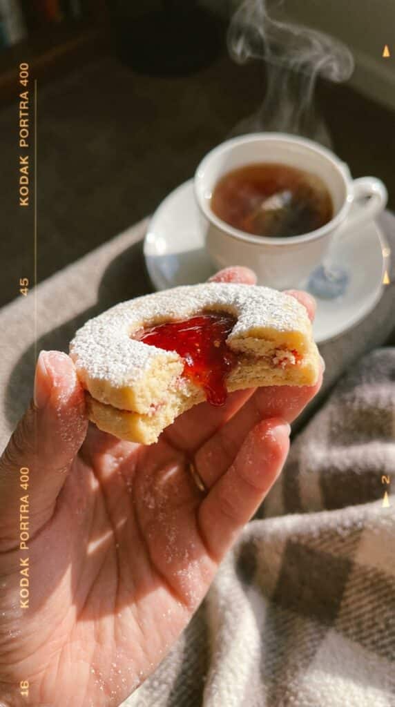 A close-up of a hand holding a heart jam cookie with a bite taken out, showing the jam filling, with a cup of tea in the background.