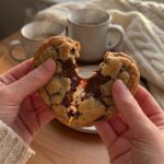 A close-up of hands breaking a warm heart-shaped chocolate chip cookie in half, showing gooey melted chocolate stretching in the middle.