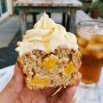 A close-up of a hand holding a halved peach cupcake, revealing the diced fruit baked inside, with frosting and a honey drip on top.
