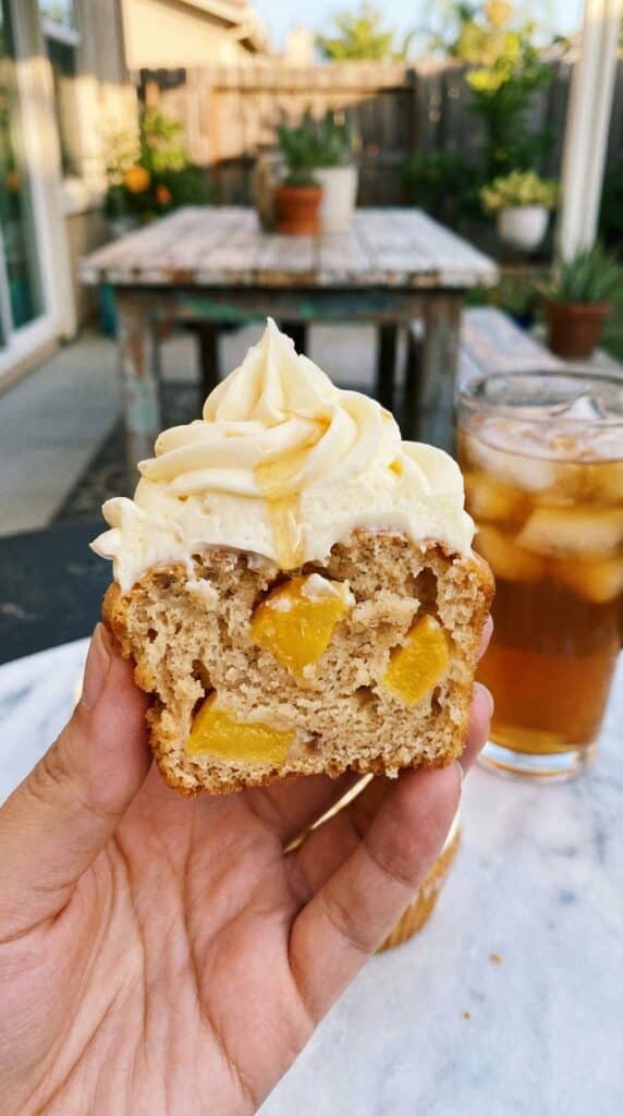 A close-up of a hand holding a halved peach cupcake, revealing the diced fruit baked inside, with frosting and a honey drip on top.