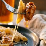A close-up of a fork holding a piece of creamy penne pasta and a bite of sticky honey pepper chicken, with a cheese pull.