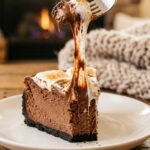 A close-up of a slice of chocolate cheesecake with a fork pulling away a bite of gooey toasted marshmallow topping.