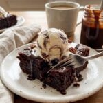 A close-up of a warm slice of brownie bread topped with melting vanilla ice cream and hot fudge drizzle, with a fork taking a bite.