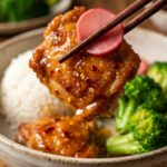 A close-up of wooden chopsticks holding a piece of glossy, sticky hot honey chicken and a pink pickled onion slice over a bowl of rice and vegetables.