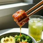 A close-up of chopsticks lifting a piece of shiny, sticky hot honey chicken from a bowl, with a drip of honey falling from it.