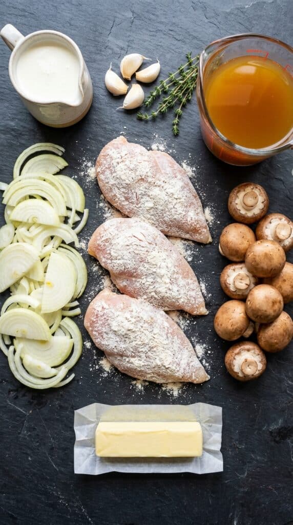 A flat lay showing raw floured chicken, sliced onions, mushrooms, chicken broth, cream, garlic, thyme, and butter on a dark slate board.