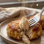 A close-up of a fork breaking into a golden crab cake, revealing huge chunks of white crab meat inside, with a lemon being squeezed in the background.