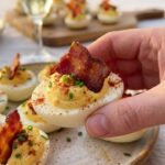 A close-up of a hand picking up a single deviled egg topped with bacon and paprika, with sparkling wine in the blurred background.
