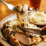 A close-up of a fork cutting into a cheesy smothered chicken breast, stretching the melted Monterey Jack cheese, with mashed potatoes in the background.