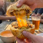 A close-up of hands pulling apart a garlic parmesan dough ball, creating a long, gooey cheddar cheese stretch, revealing ground beef inside.