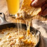 A close-up of a tortilla chip scooping up a thick, cheesy bite of hot crab dip with a cheese pull.